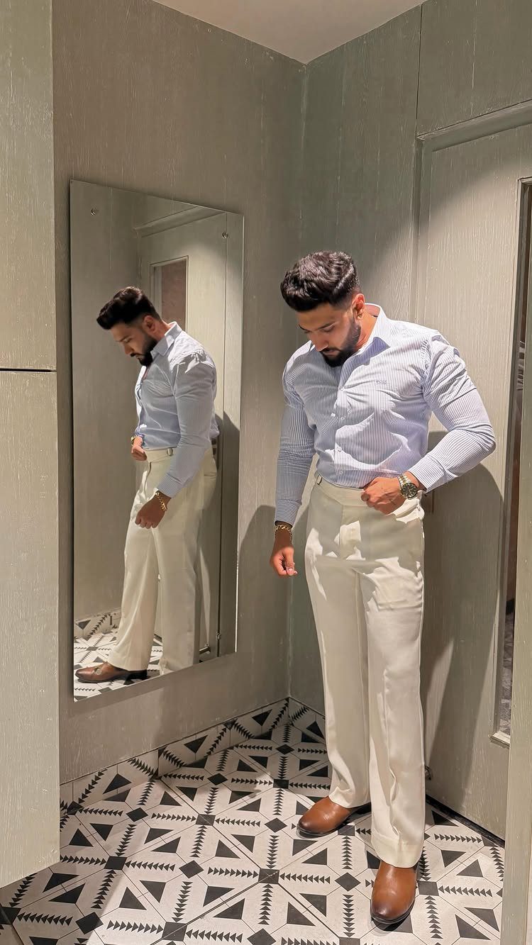 Man wearing a striped blue shirt, cream trousers, and brown leather shoes standing near a mirror.