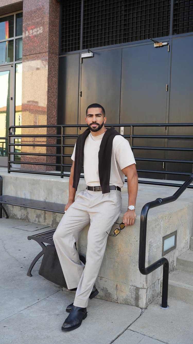 A man in cream t-shirt and beige trousers posing outdoors against a dark building.