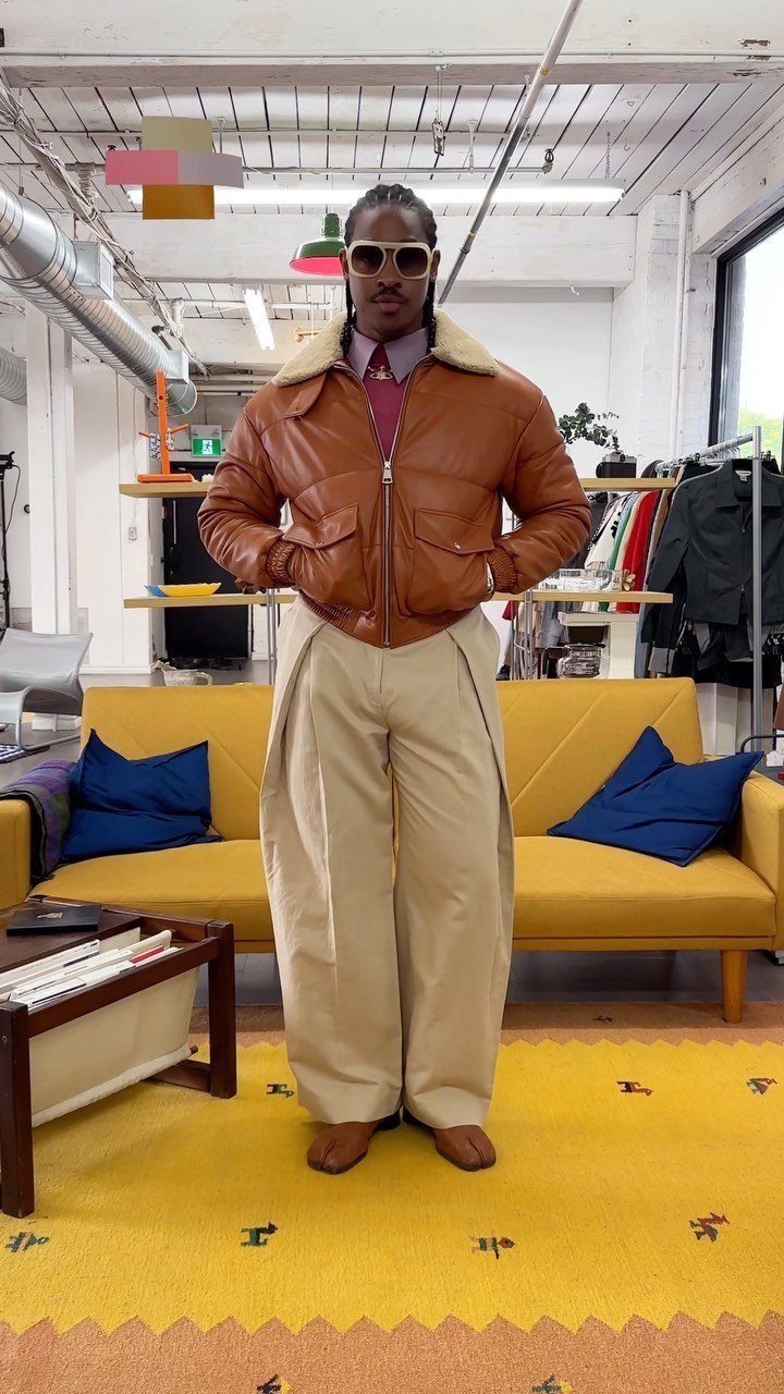 A man posing in a brown leather jacket, maroon shirt, and wide-leg beige trousers.