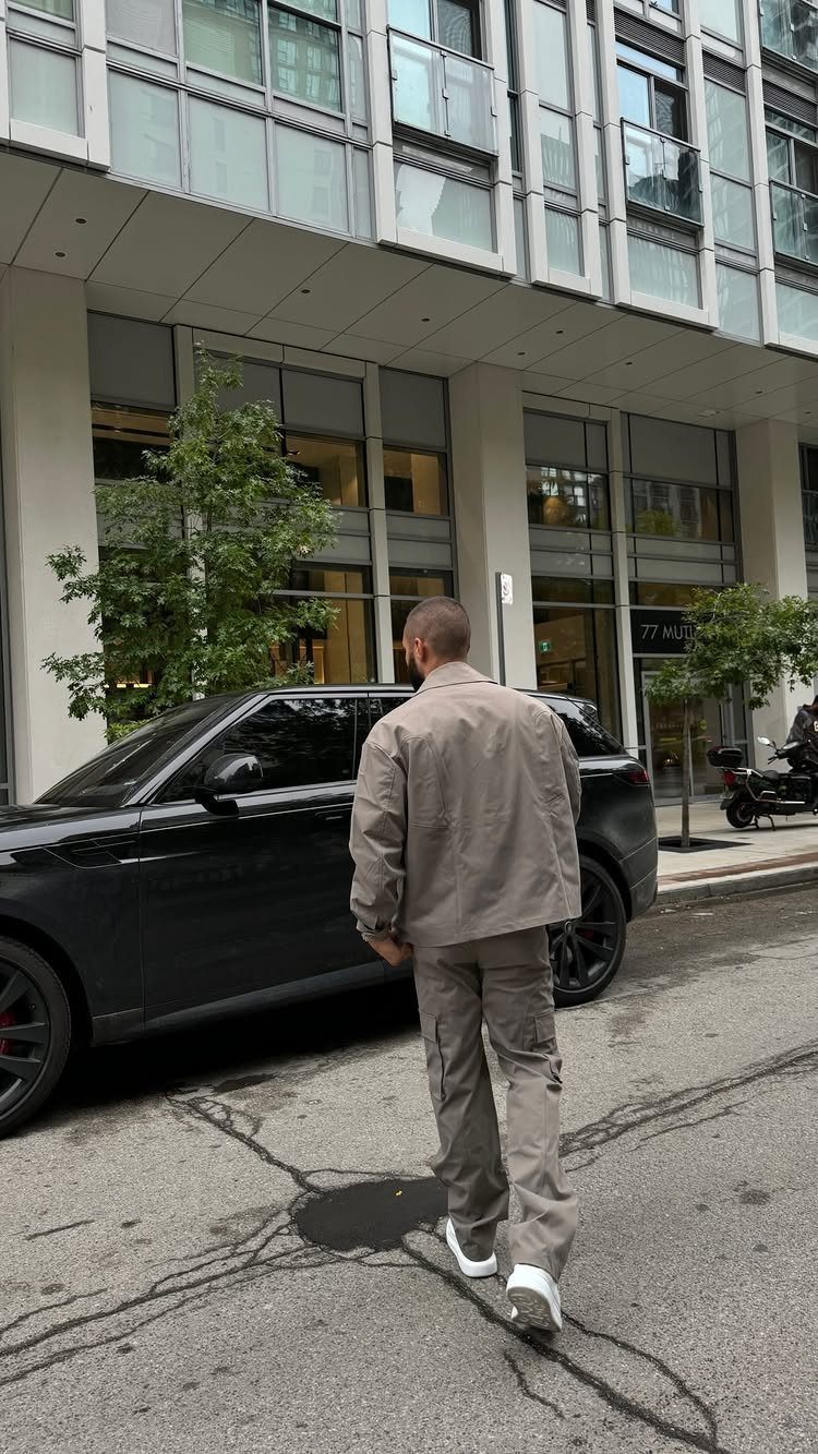 A man in a tan jacket and cargo pants walking past a black car on a city street.