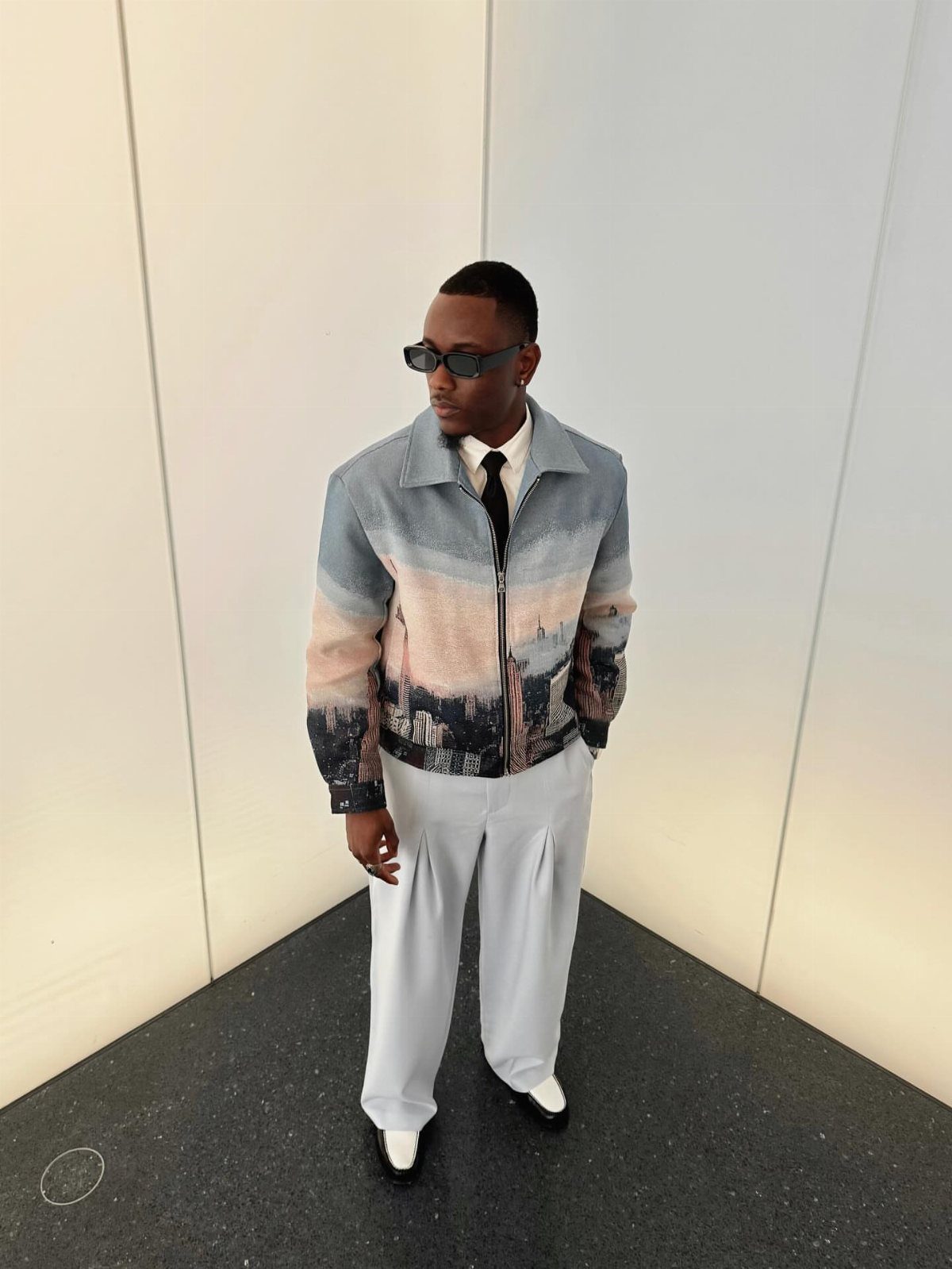 A man wearing a landscape print jacket, black tie, and light grey trousers standing in a corner.