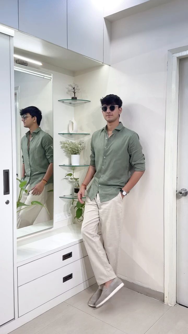 A man wearing a sage green shirt, beige trousers, and sunglasses posing against a white wall.