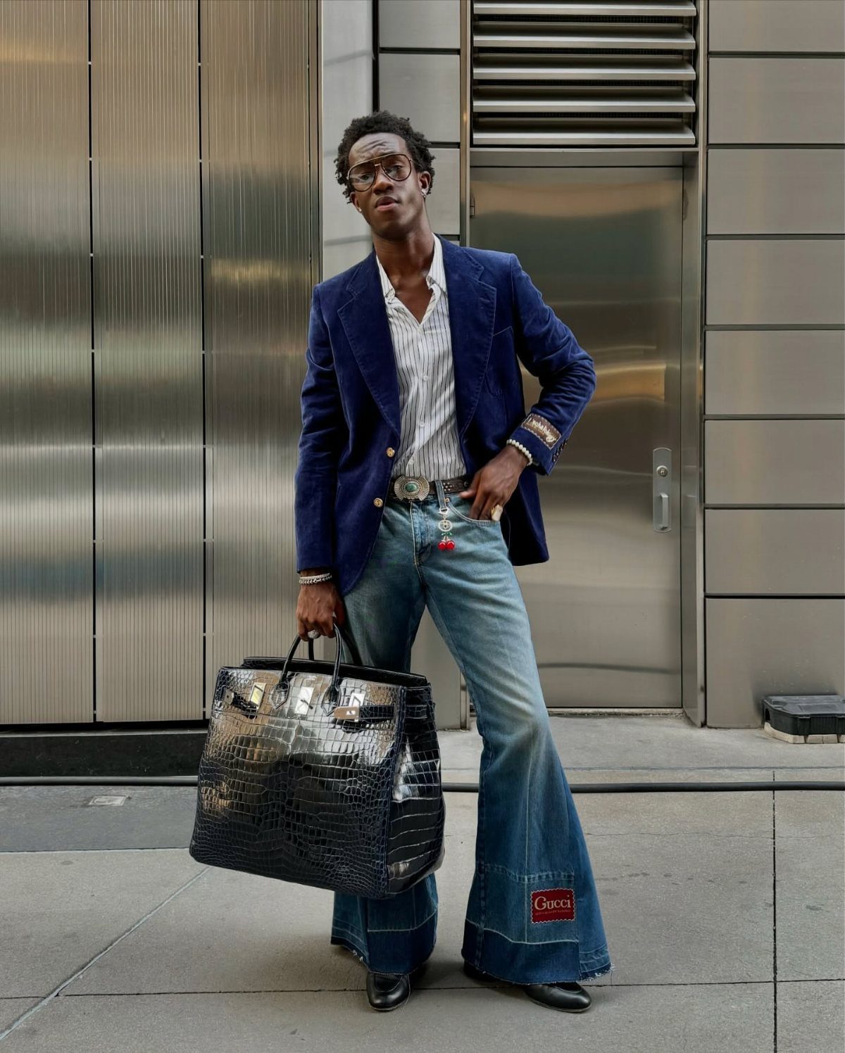 Man in blue velvet blazer and flared jeans holding a large black crocodile-patterned bag.