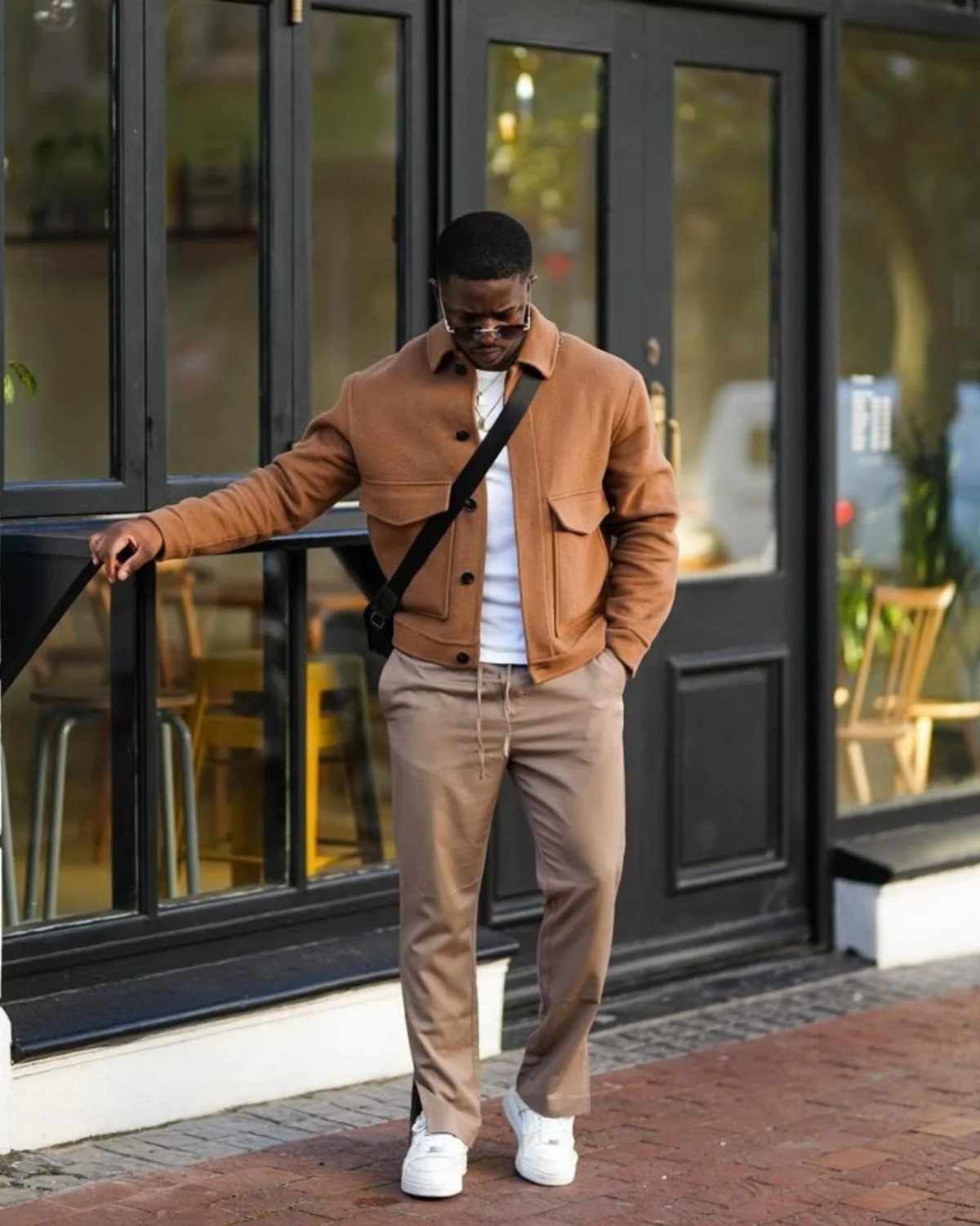 Man in a brown utility jacket, tan trousers, and white sneakers standing by a glass door.