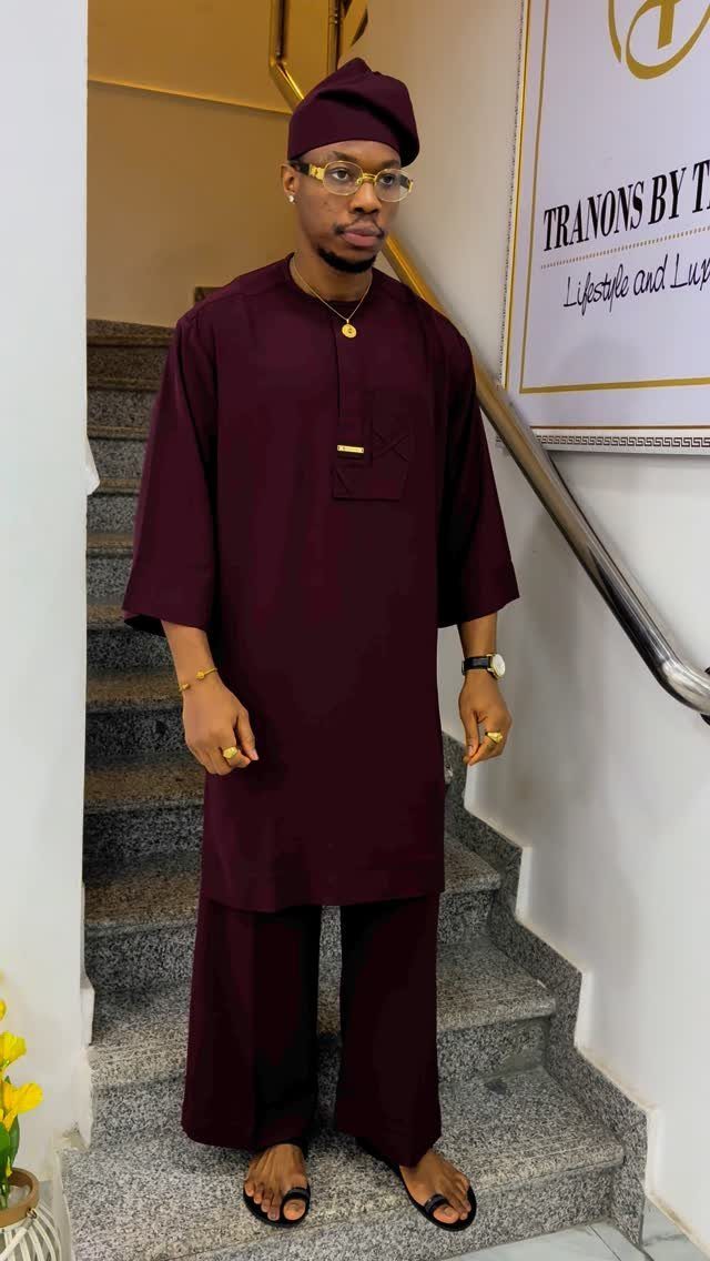 Man in burgundy traditional African attire standing on a stone staircase wearing gold accessories.