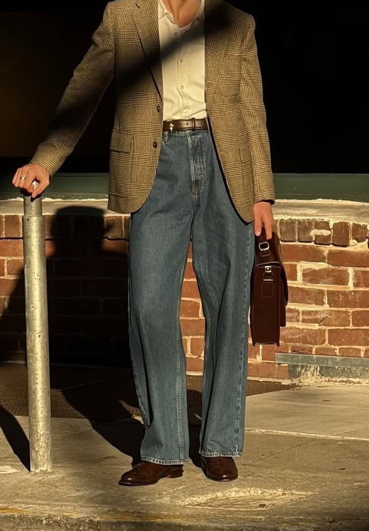 Man wearing a plaid blazer, white shirt, wide-leg jeans, and brown leather shoes holding a briefcase.