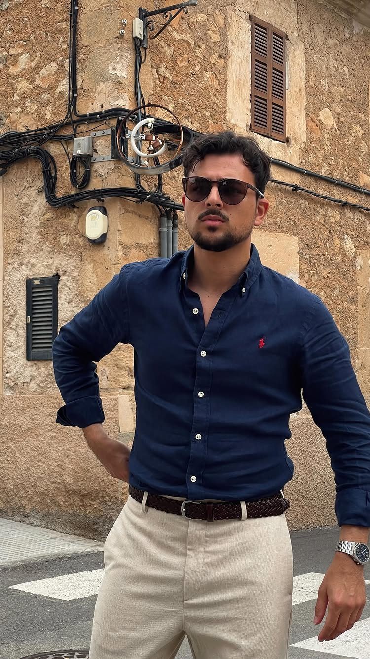 Man in navy blue shirt and beige trousers posing against a stone wall.