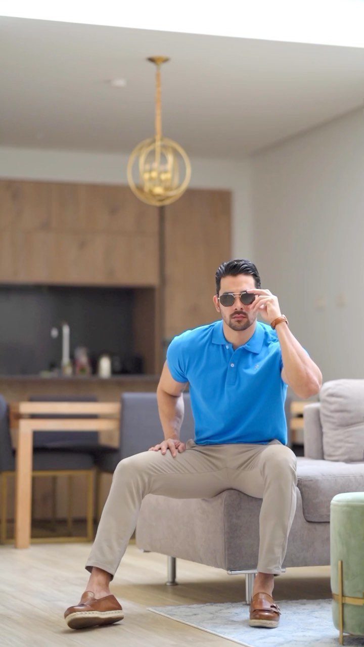 Man in a blue polo shirt and beige trousers sitting on a sofa wearing sunglasses.