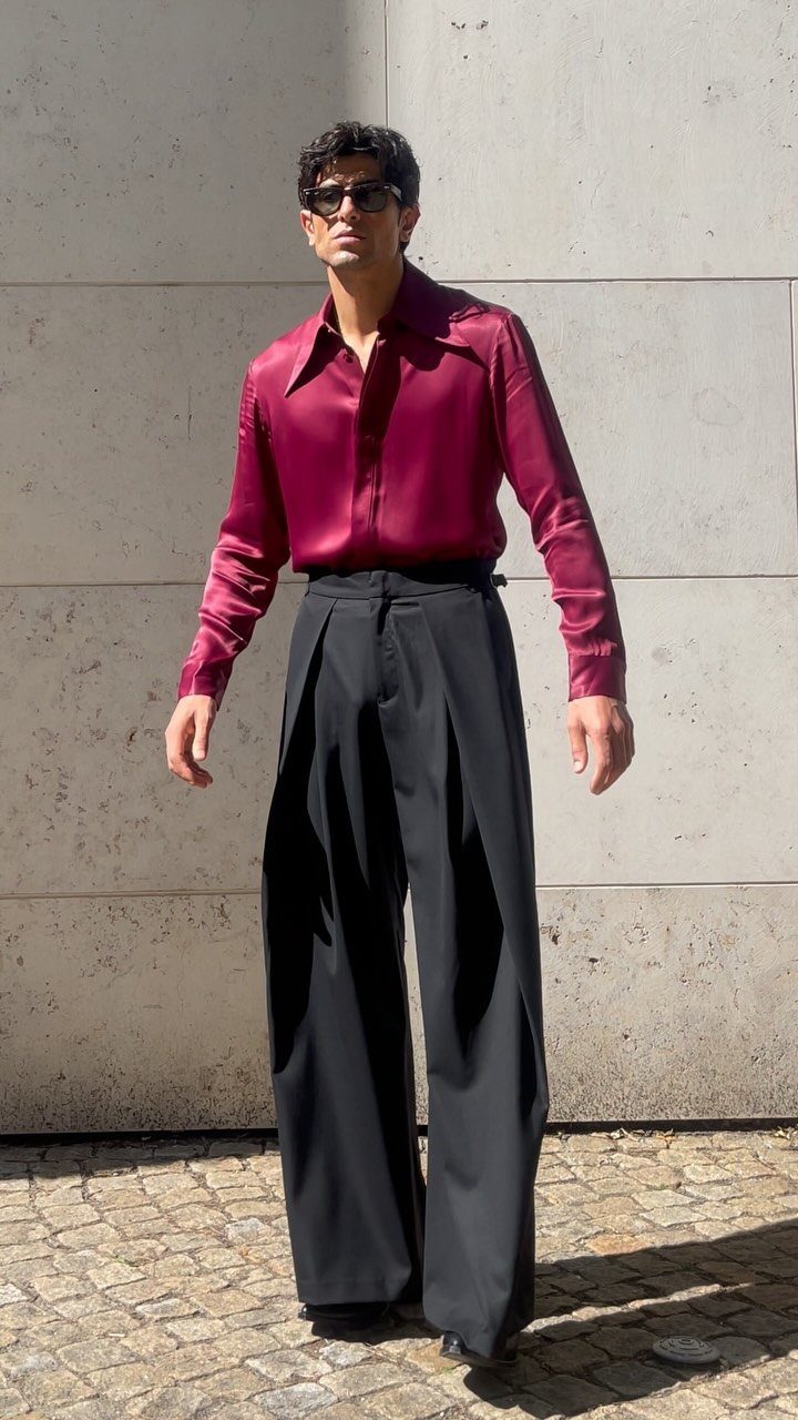 Man wearing a shiny burgundy shirt and wide-leg black trousers standing against a stone wall.