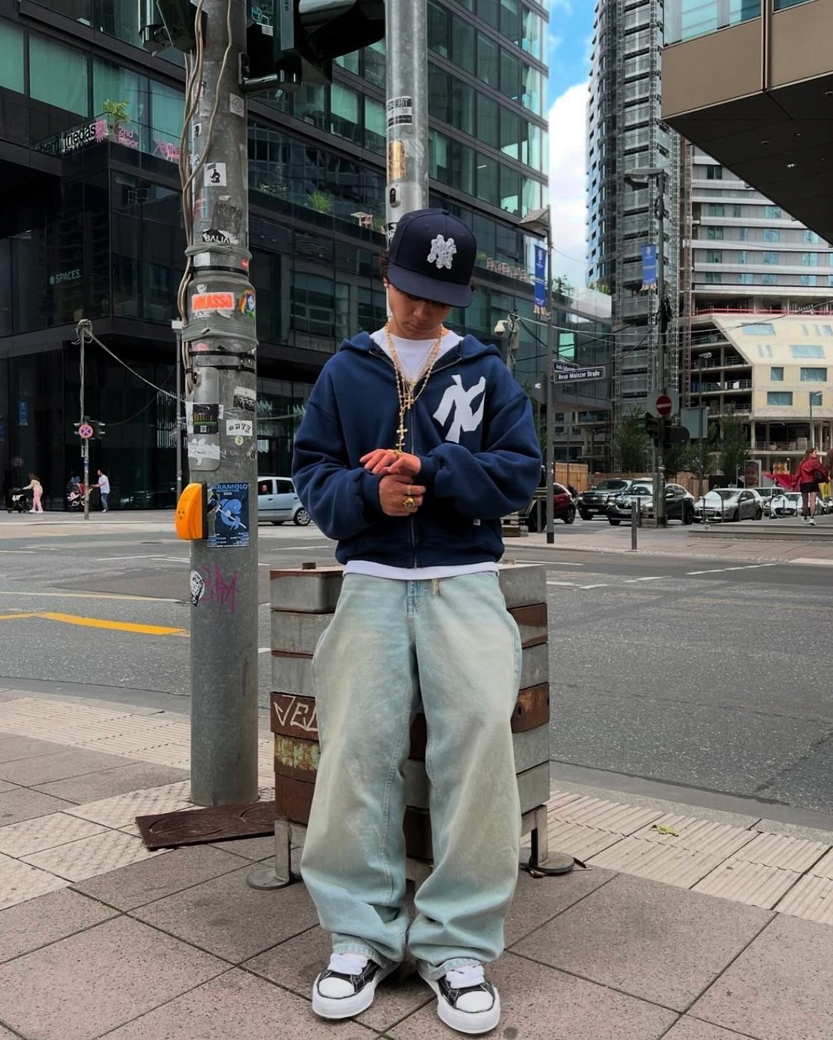 Young man wearing a navy Yankees hoodie, baggy jeans, and a baseball cap on a city street.