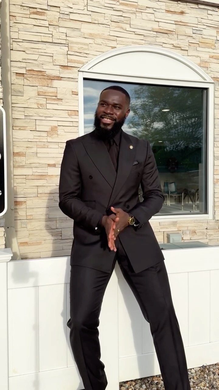 A bearded man smiling while wearing a formal all-black double-breasted suit outdoors.