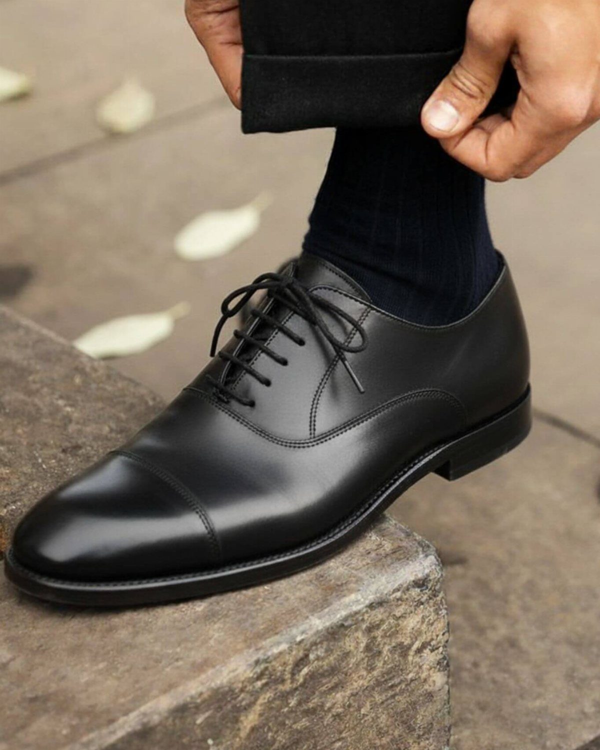 Black leather oxford shoes worn with black socks and trousers on a stone step.