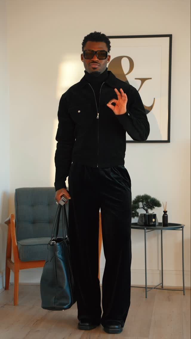 A man posing in an all-black outfit with a jacket, turtleneck, and wide-leg trousers.