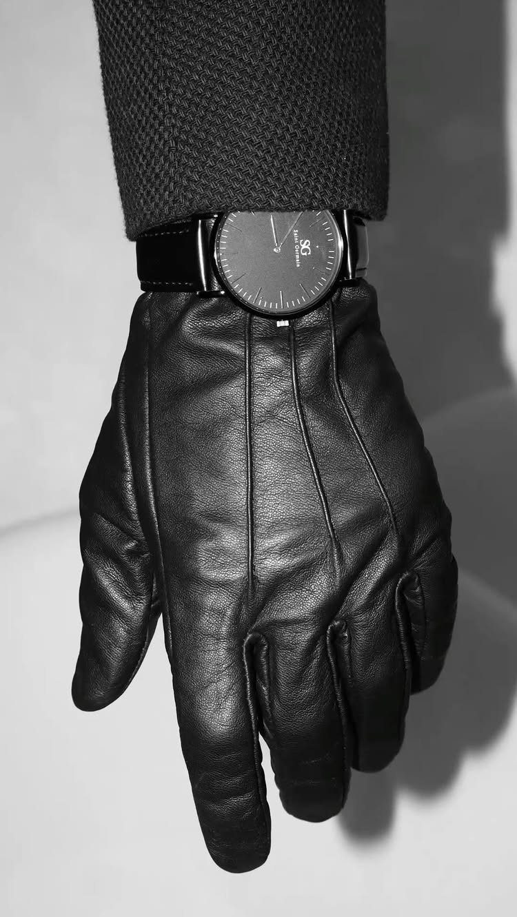 Black leather glove and a minimalist black wristwatch worn on a hand against a light background.