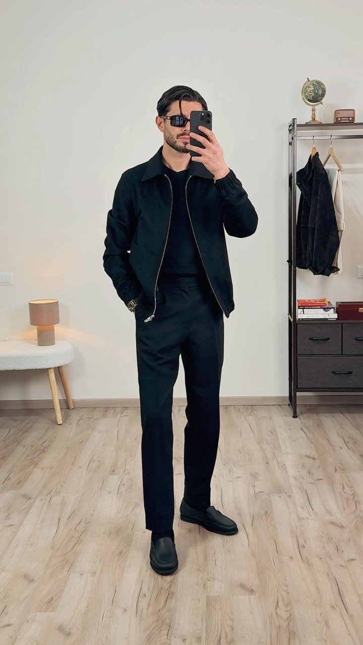 A man in an all-black outfit posing for a mirror selfie in a room.