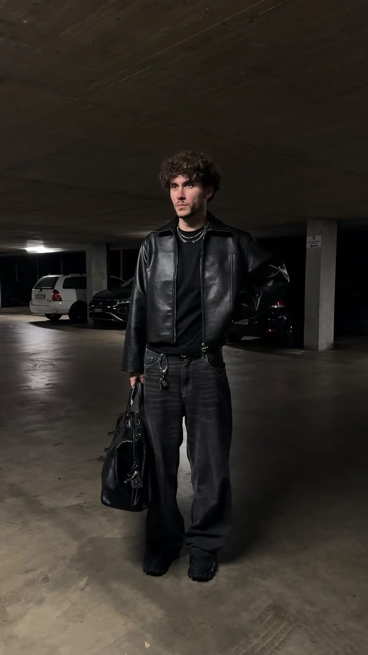 Man in an all-black outfit featuring a leather jacket standing in a dark parking garage.