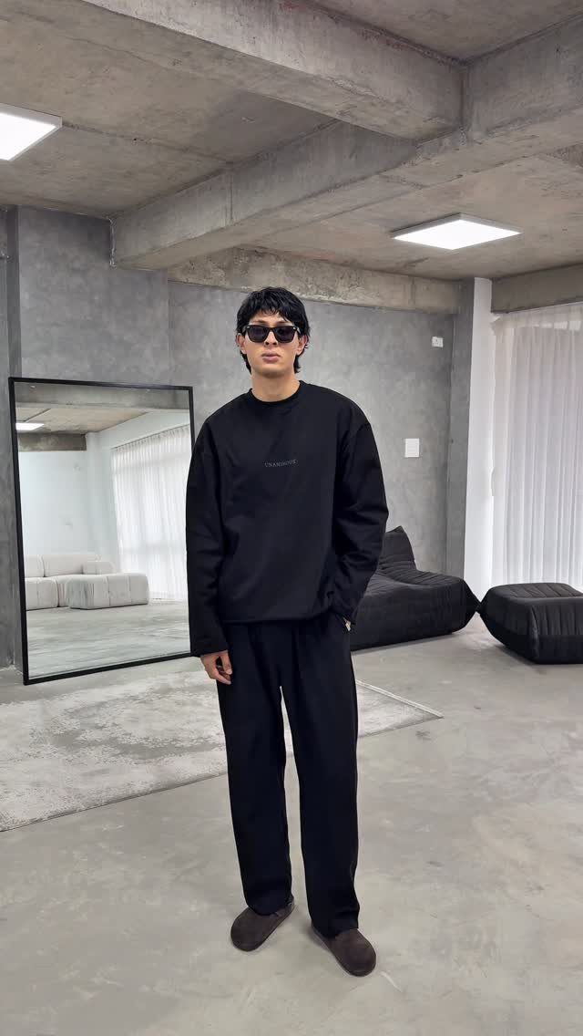 Man in oversized black top and tapered trousers standing in a concrete industrial room.
