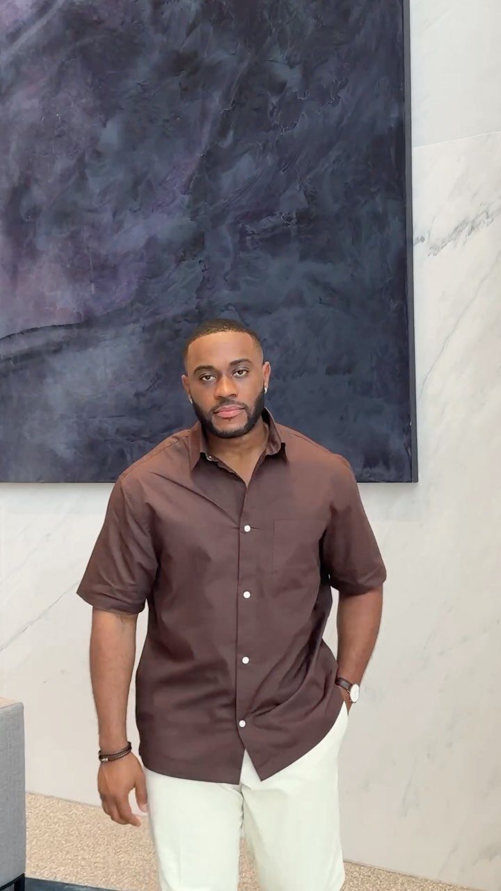 A man wearing a brown short-sleeved button-up shirt and white pants standing indoors.