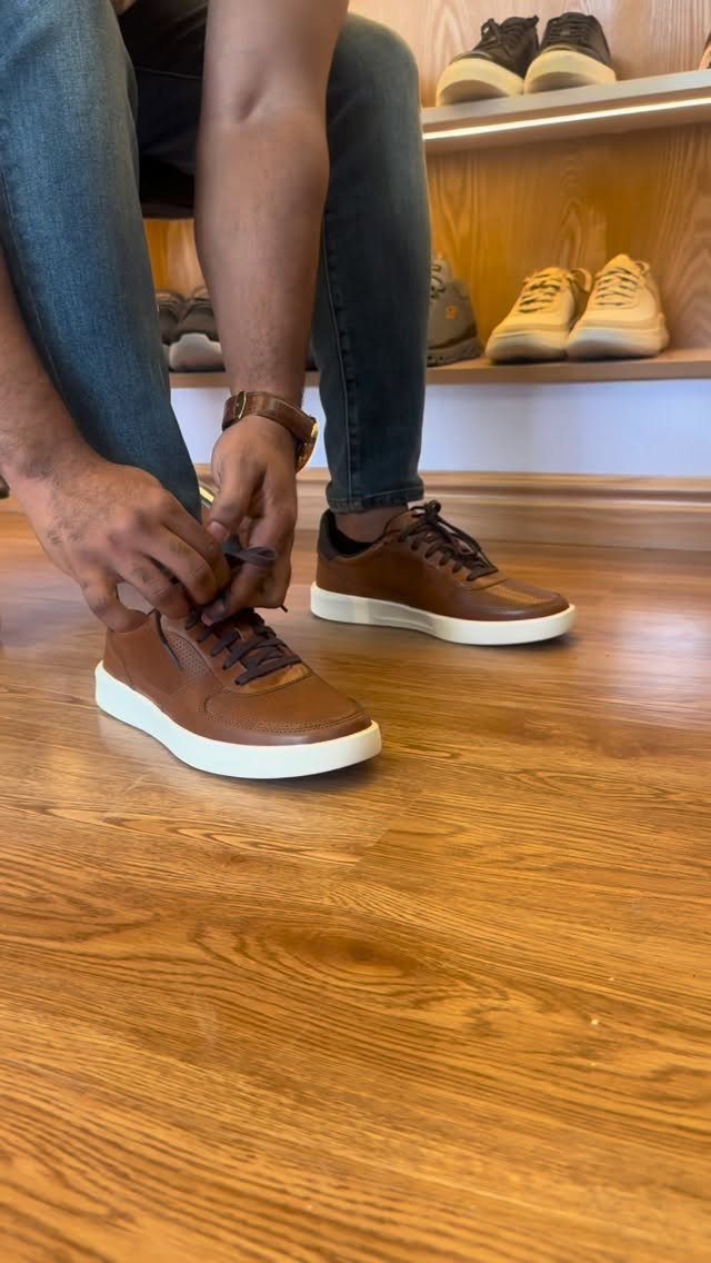 A person tying the laces of brown leather casual shoes with white soles on a wooden floor.