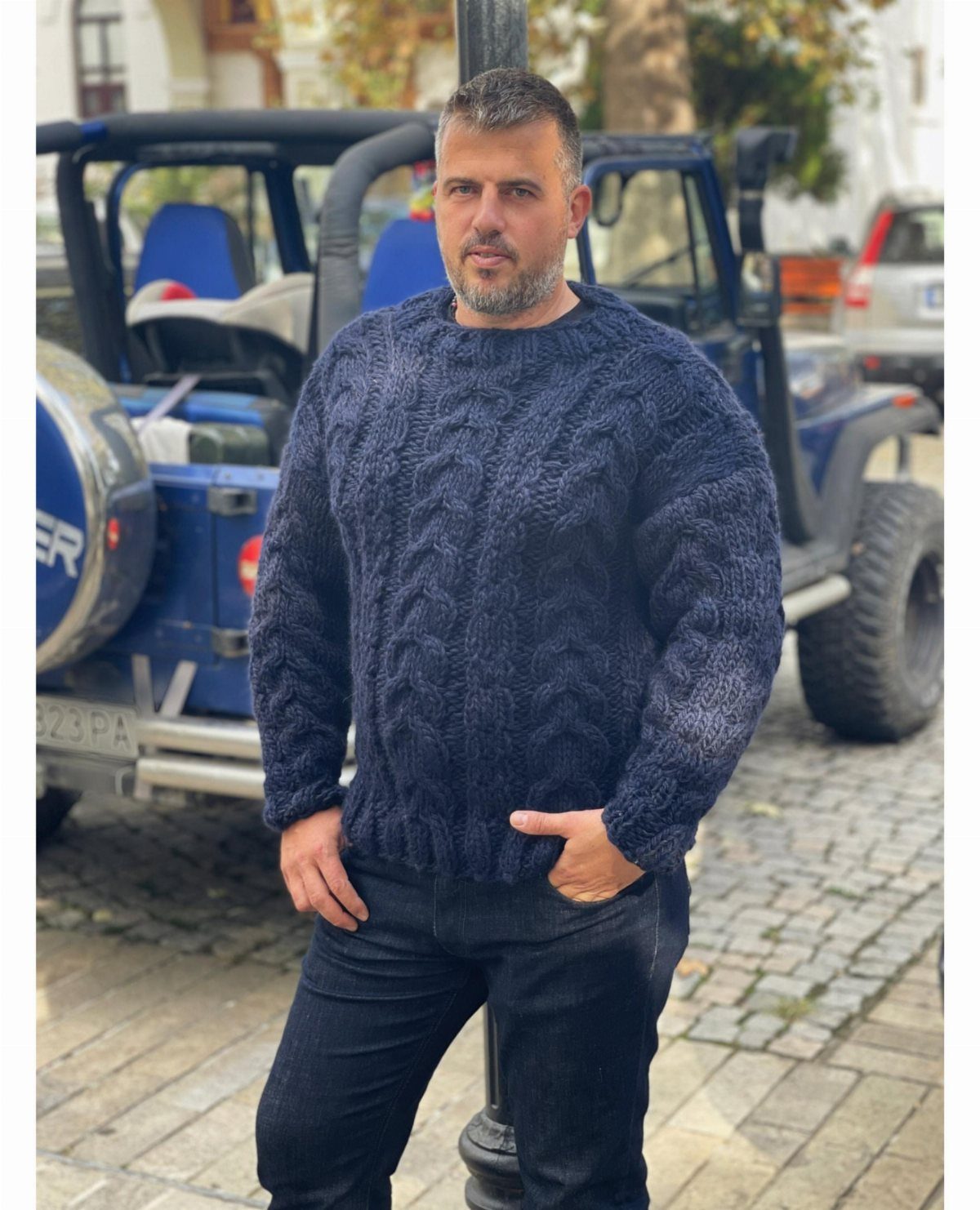 A man wearing a navy blue cable-knit crew neck sweater and dark jeans standing outdoors.
