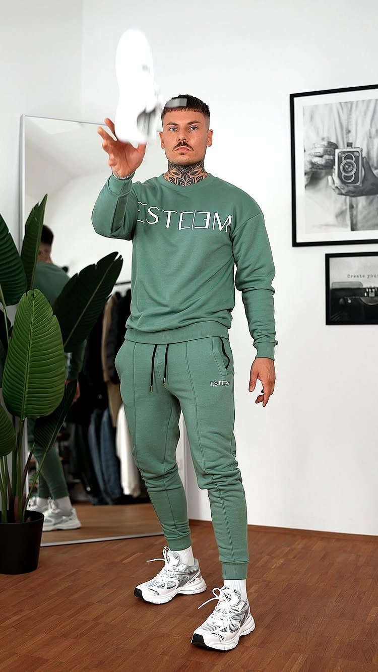 Man in sage green sweatshirt and matching joggers holding up a white sneaker.