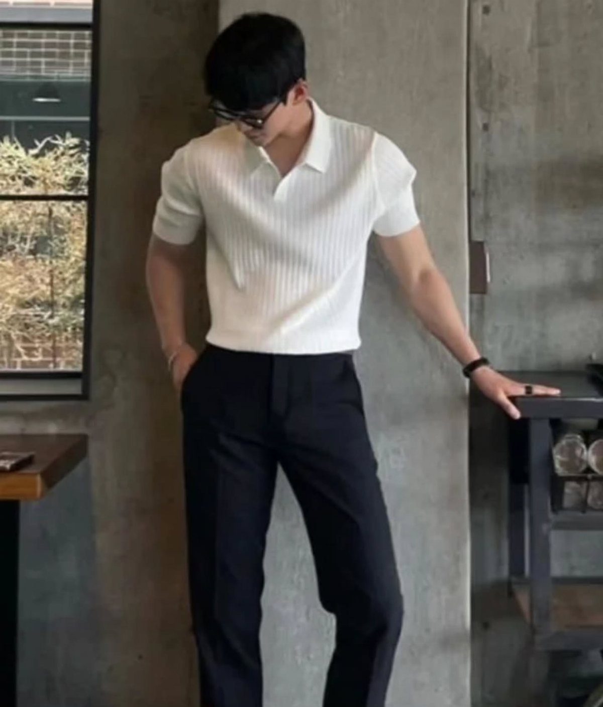 A man in a white textured polo shirt and black trousers leaning against a wall.