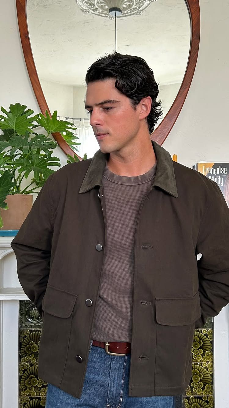 Man wearing a dark brown utility jacket with a green collar over a brown sweater and blue jeans.