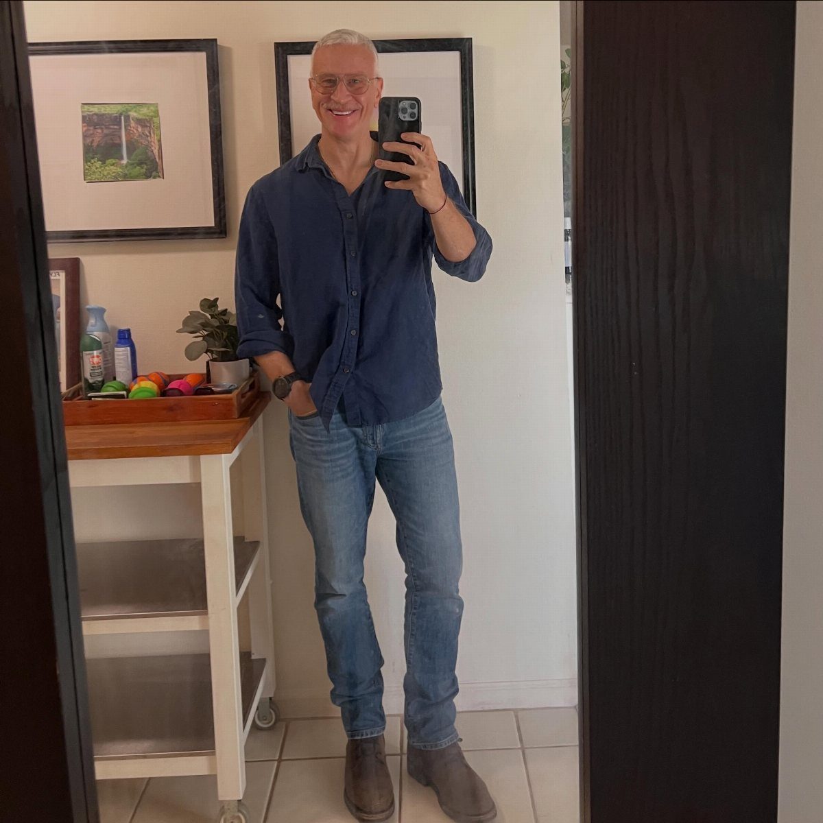 A man in a blue button-down shirt and denim jeans taking a mirror selfie.