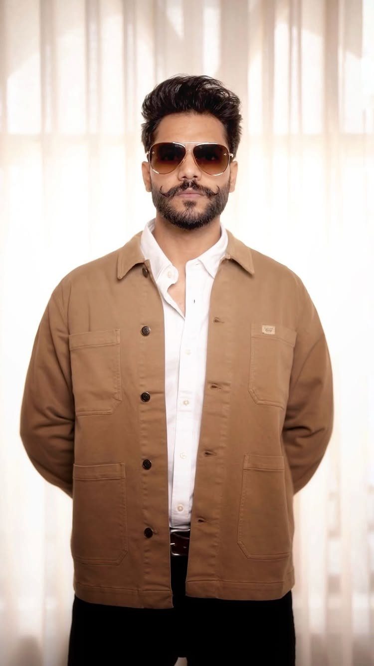 Man wearing a tan utility jacket over a white button-down shirt and sunglasses.