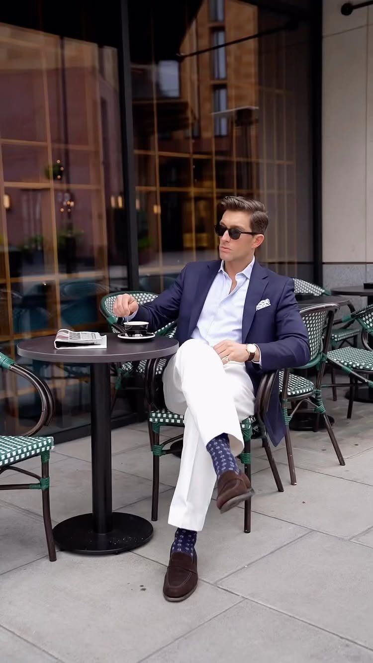 Man in navy blazer and white trousers sitting at a cafe wearing brown suede dress shoes.