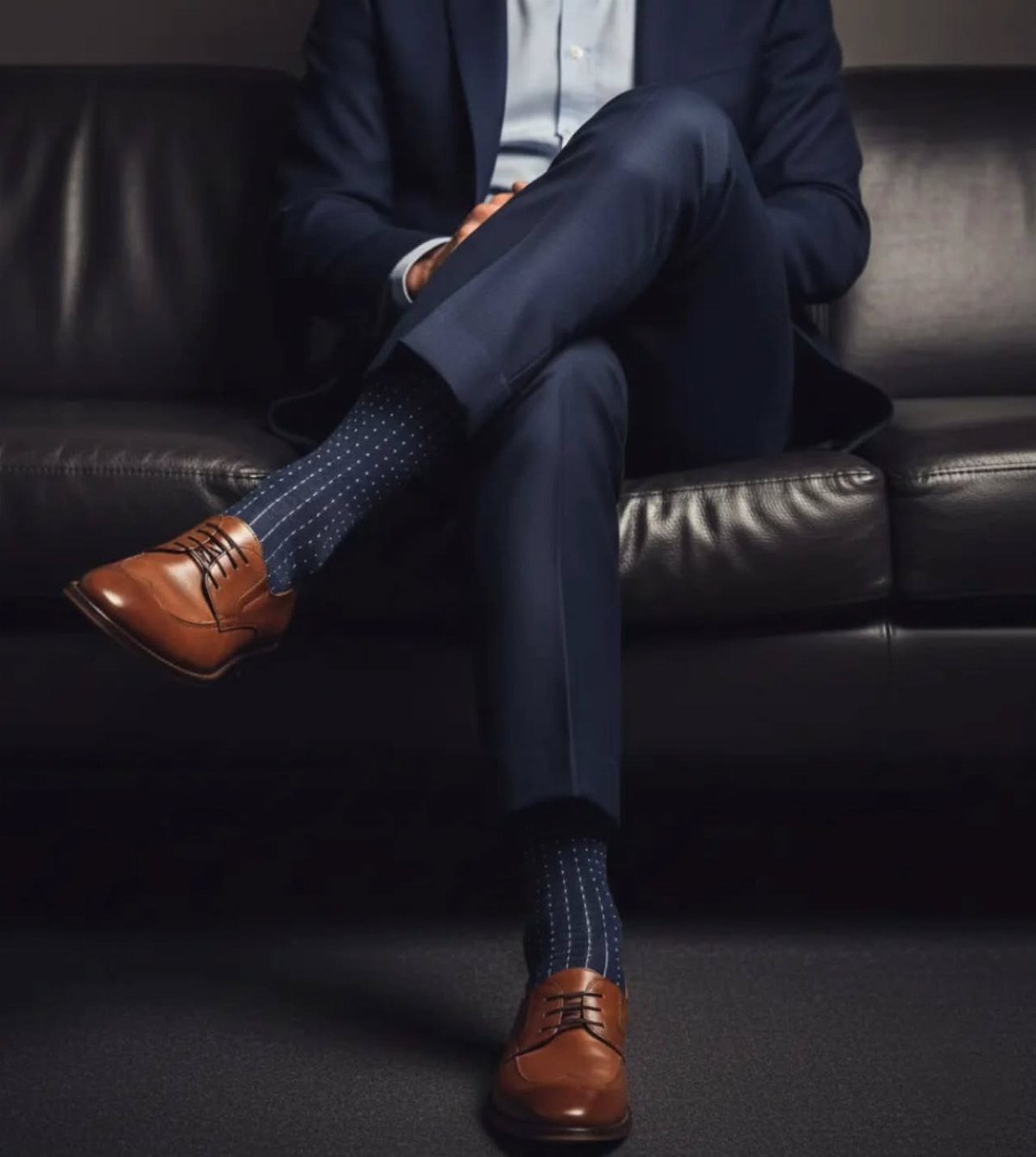 Man in a navy suit sitting on a leather sofa wearing polka dot crew socks and brown shoes.
