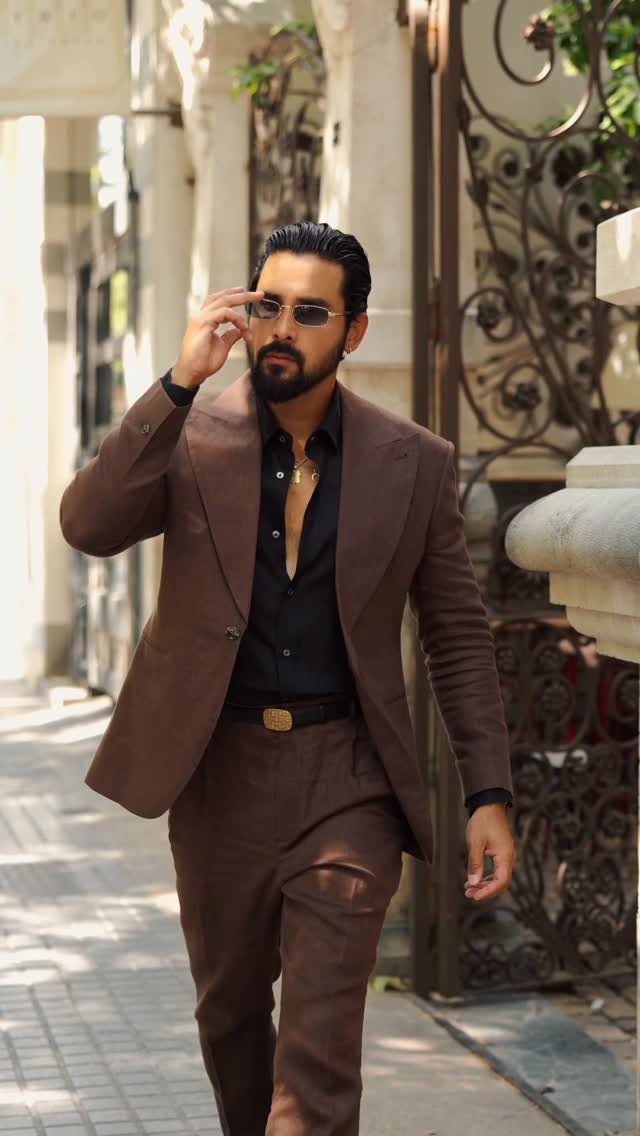 Man in a well-fitted brown suit and black shirt walking down a sunny street.