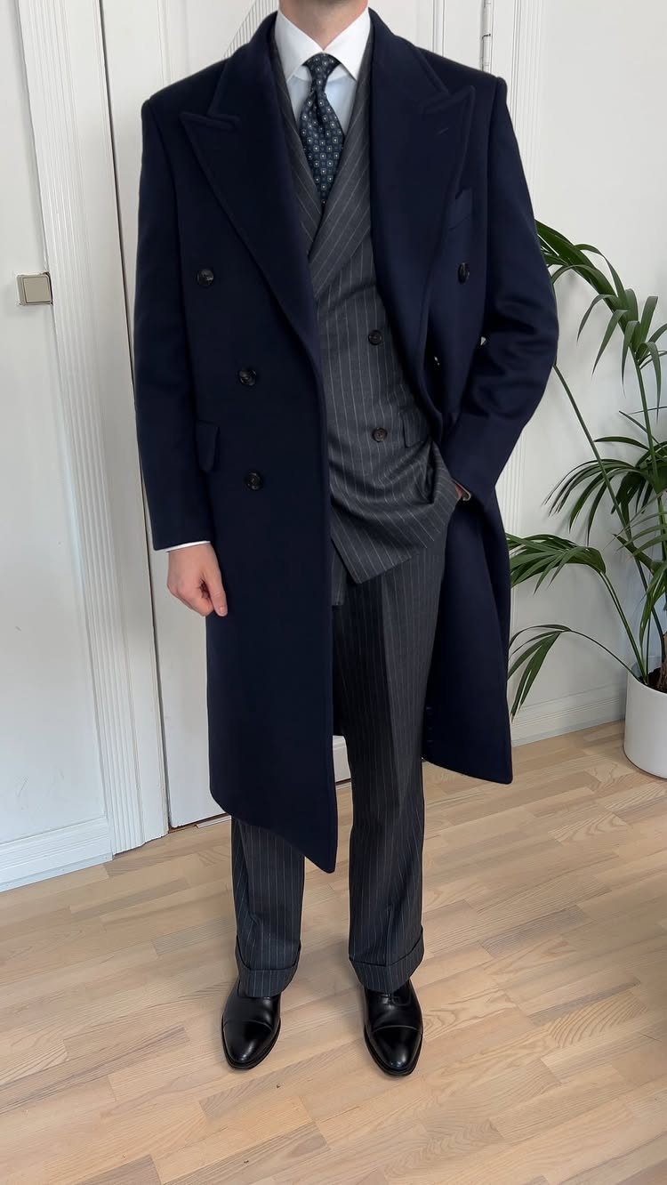 Man wearing a navy overcoat over a grey pinstripe suit and tie.