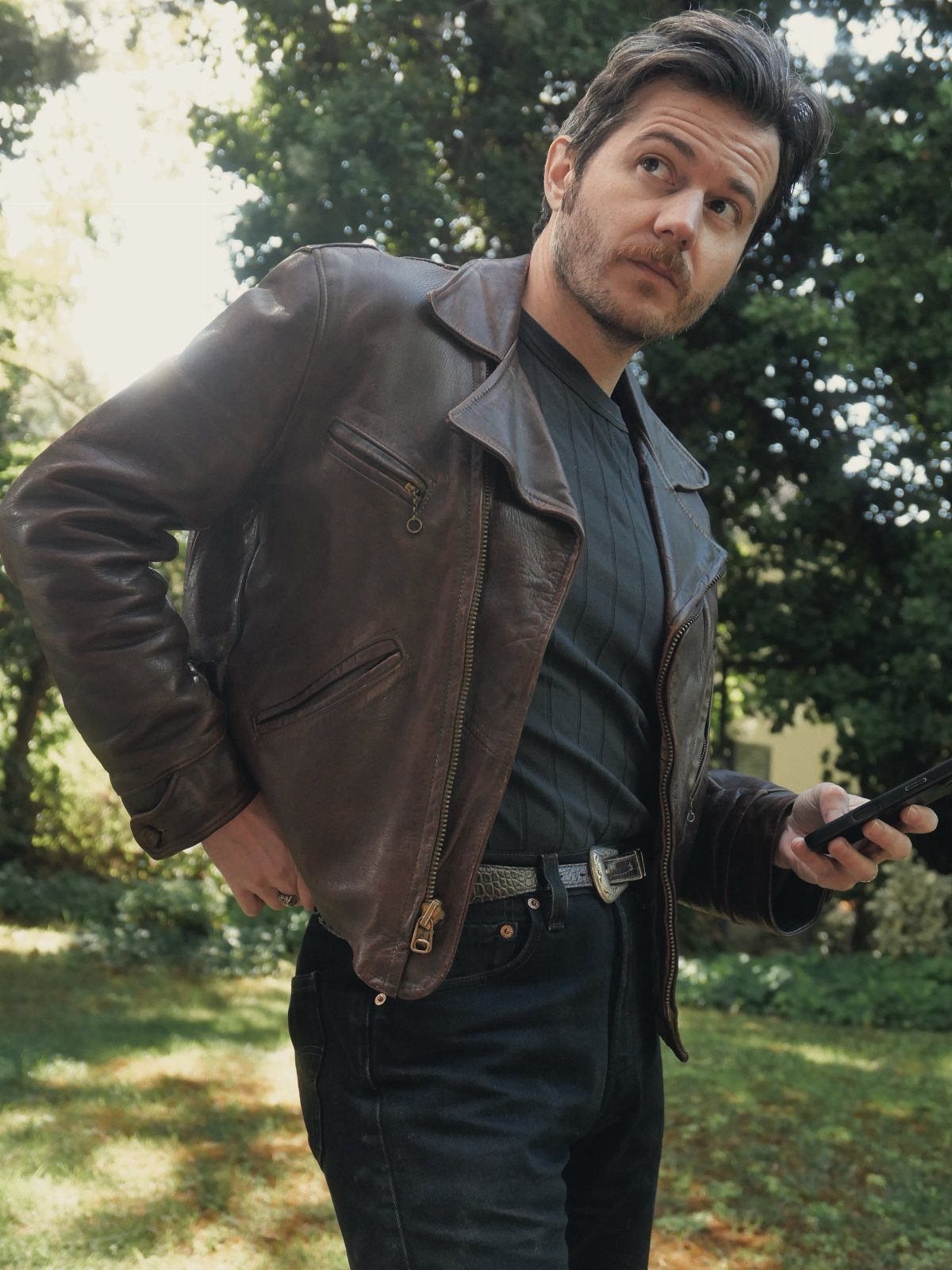 Man in a brown leather jacket and black denim jeans standing in a sunlit garden.