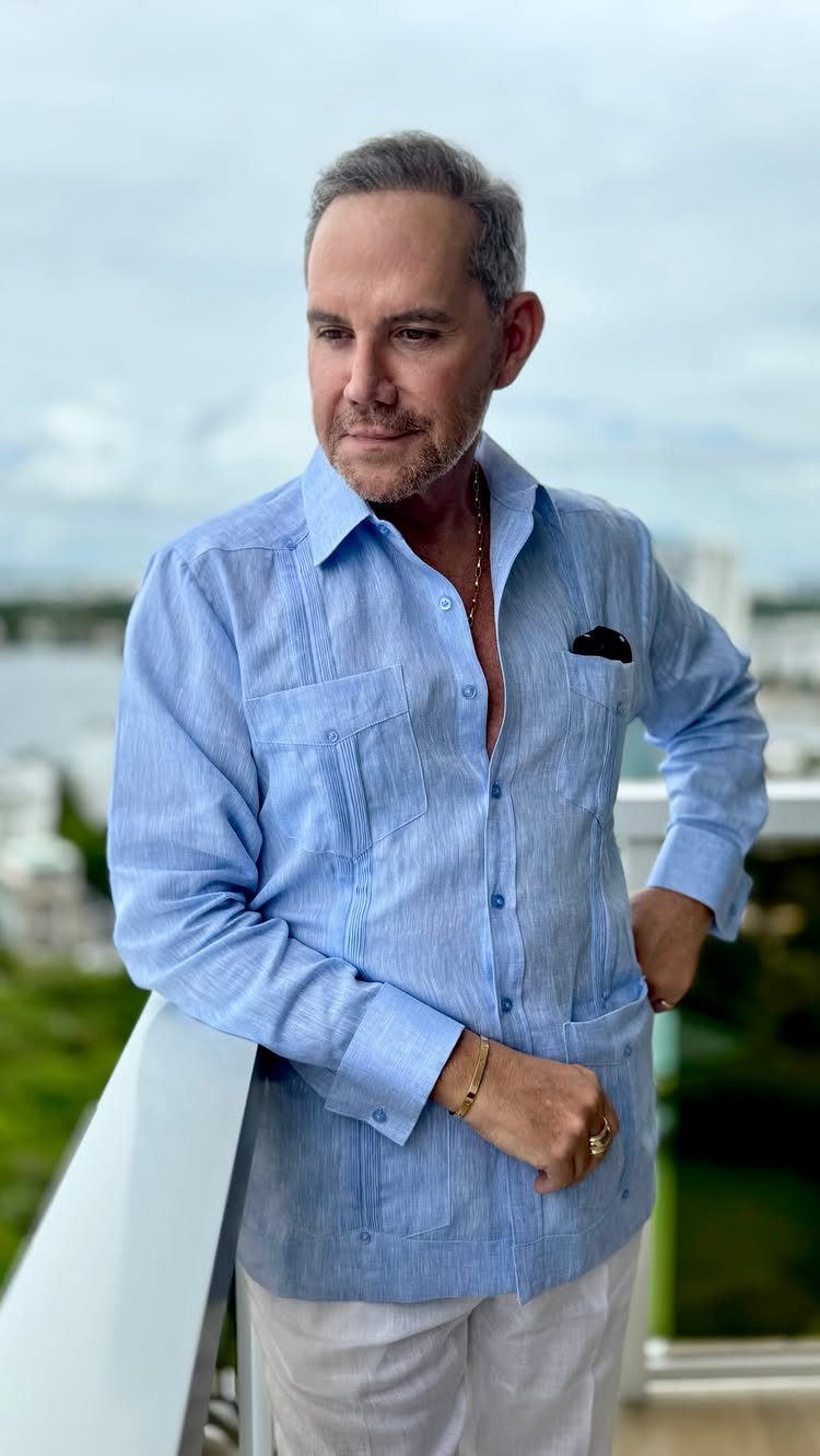 Man in a light blue linen shirt standing on a balcony overlooking a coastal city landscape.