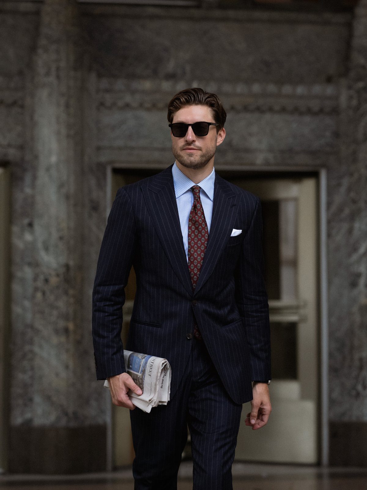 Man in a dark pinstripe business suit and sunglasses holding a newspaper outdoors.