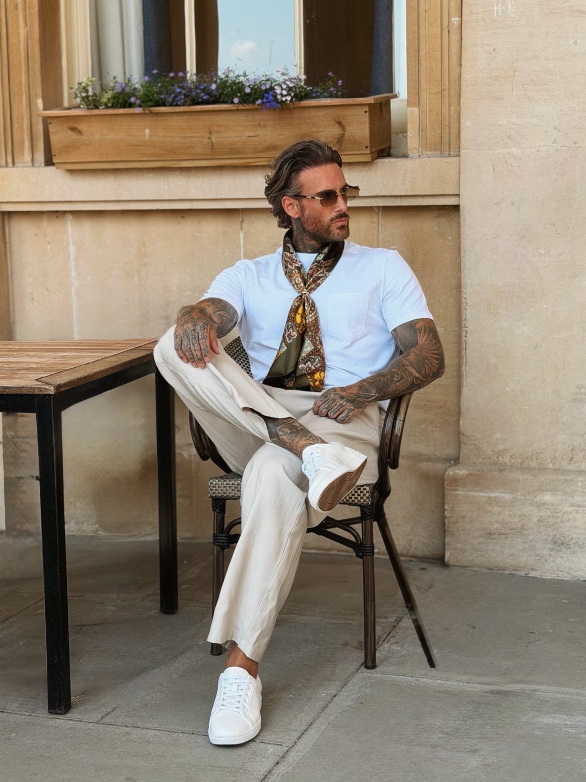 Man in white t-shirt, beige trousers, and a patterned neck scarf sitting outdoors.