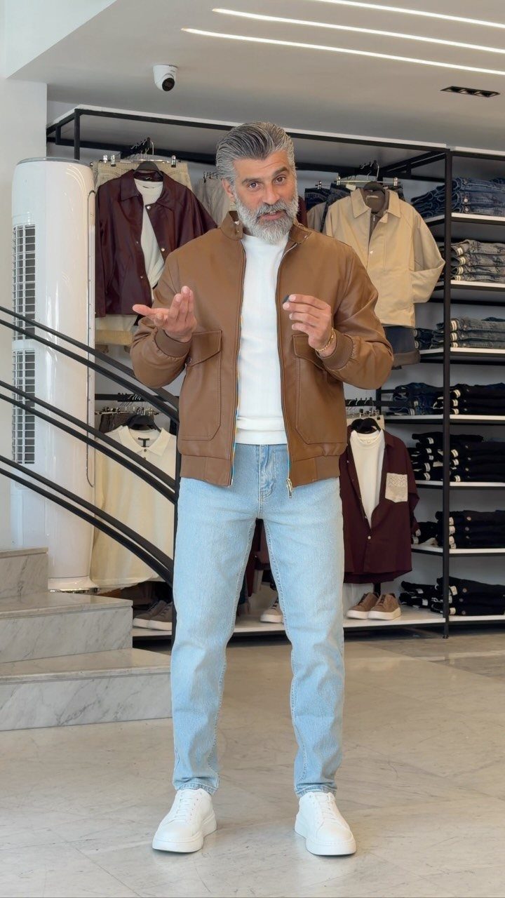 Man with grey hair wearing a brown leather jacket, white sweater, light blue jeans, and white sneakers.