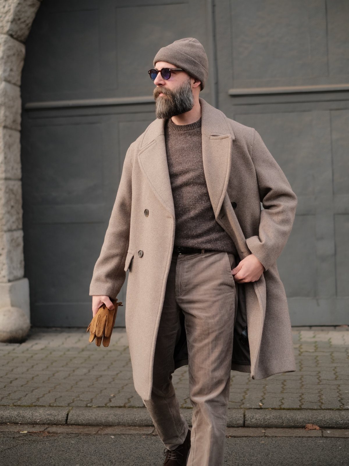 Bearded man walking in a monochromatic tan wool overcoat, sweater, and corduroy trousers.