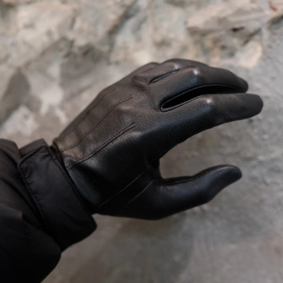 Black leather gloves worn on a hand against a textured grey stone background.