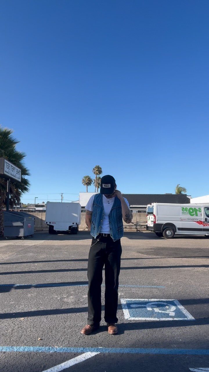 Man wearing a blue denim vest, white t-shirt, and black trousers standing in a parking lot.