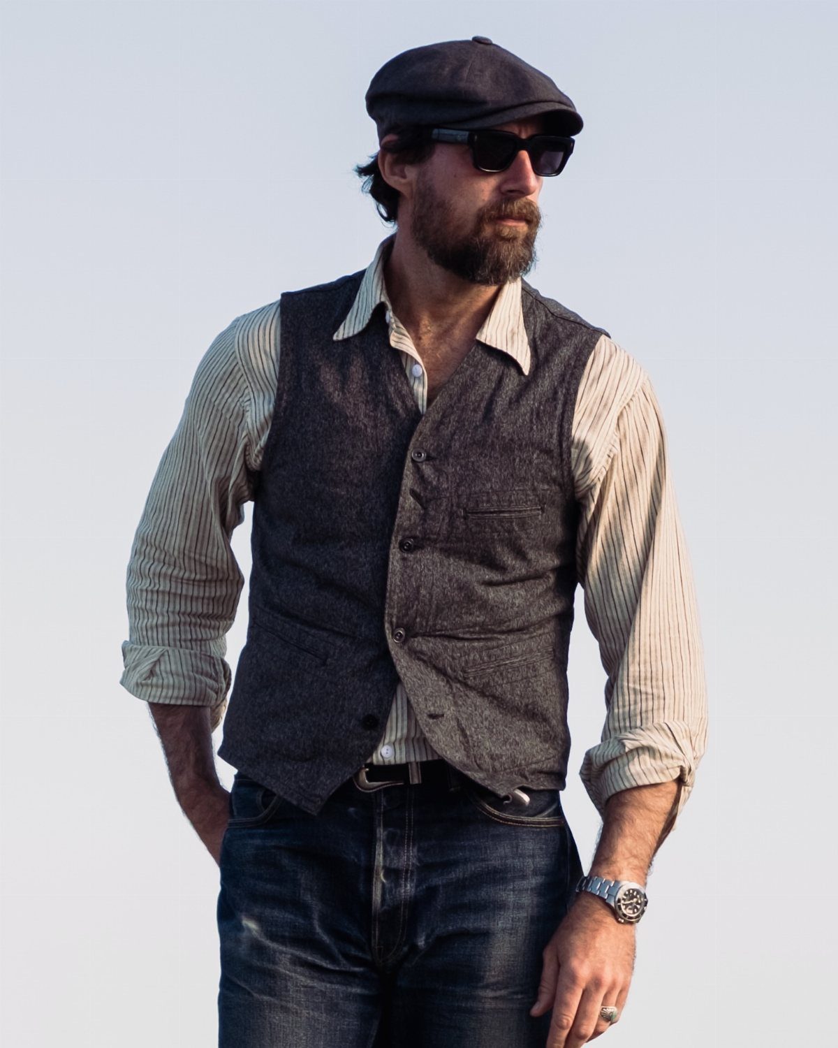 A man wearing a grey vest, striped shirt, and blue jeans standing outdoors.