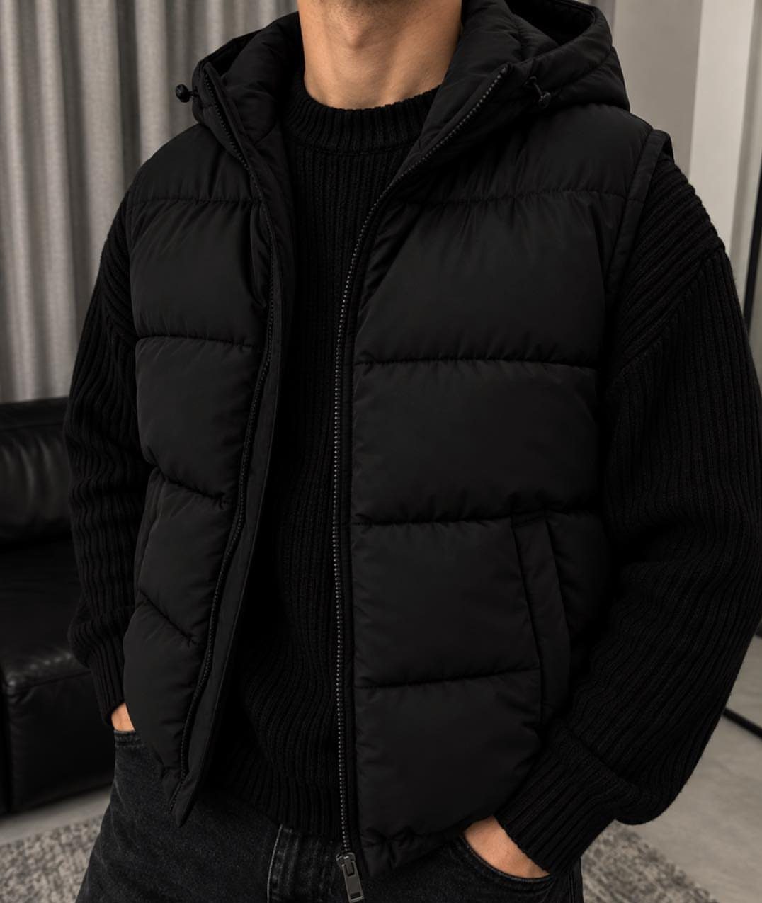 A man wearing a black quilted puffer vest over a black ribbed sweater and dark jeans.