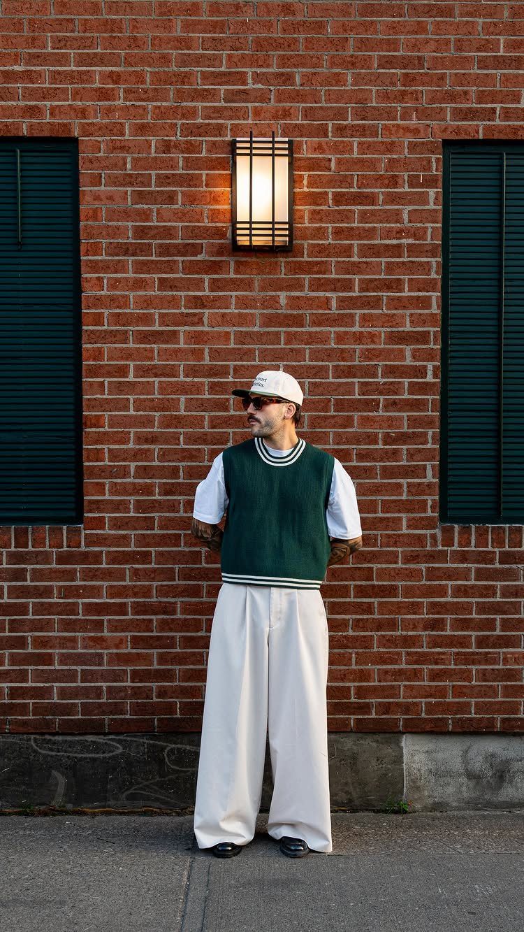 Man in a dark green sweater vest, white t-shirt, and wide cream trousers standing by a brick wall.