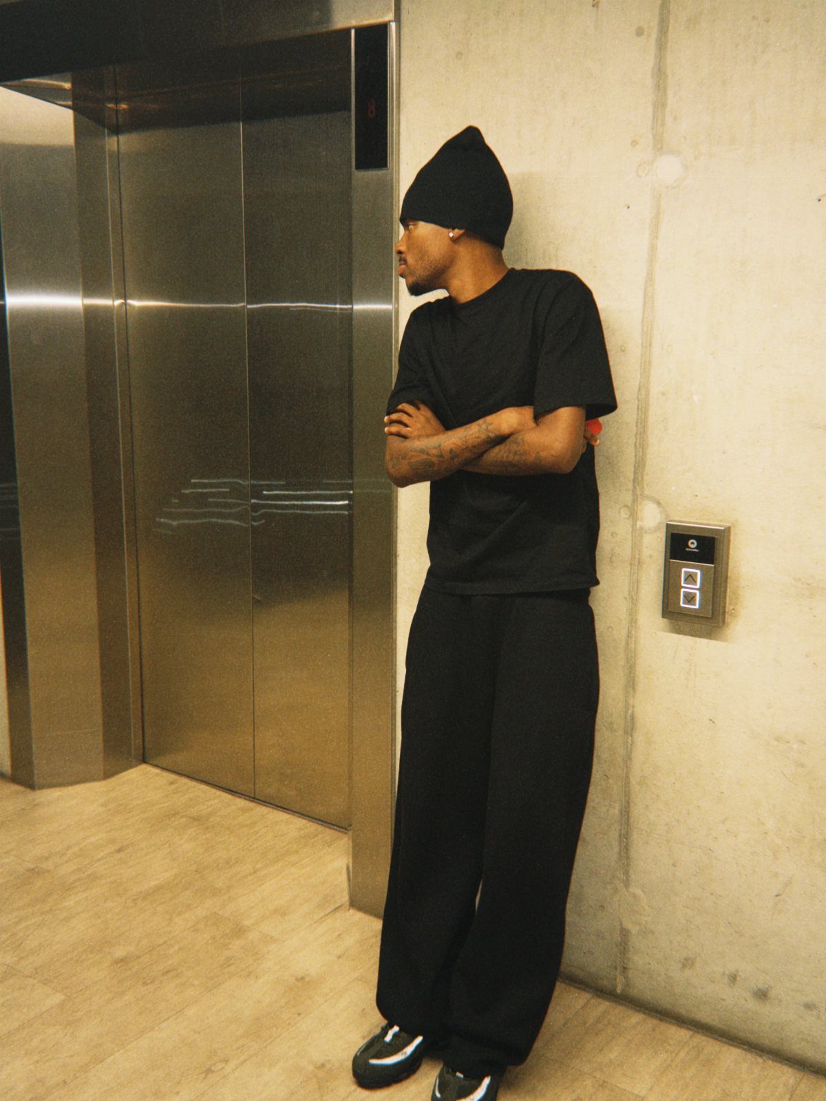 Man in an all-black outfit wearing a beanie, t-shirt, and wide-leg sweatpants by an elevator.