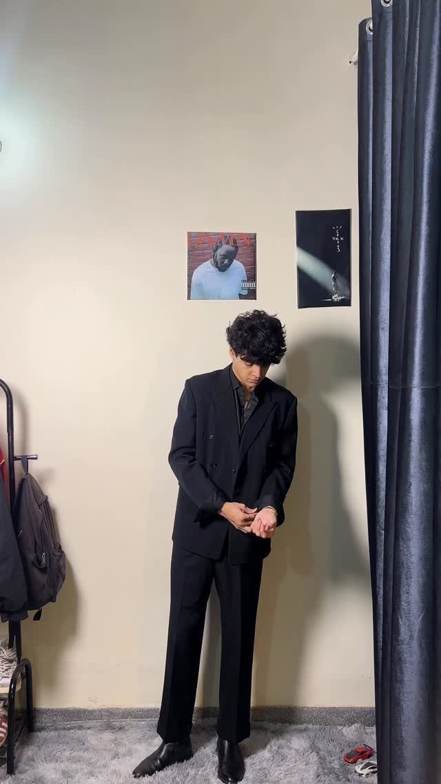 Man in an all-black blazer, shirt, and trousers standing in a room.