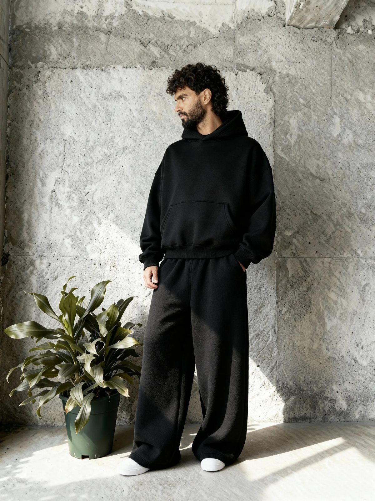 Man in a black hoodie and wide-leg black sweatpants standing by a potted plant.