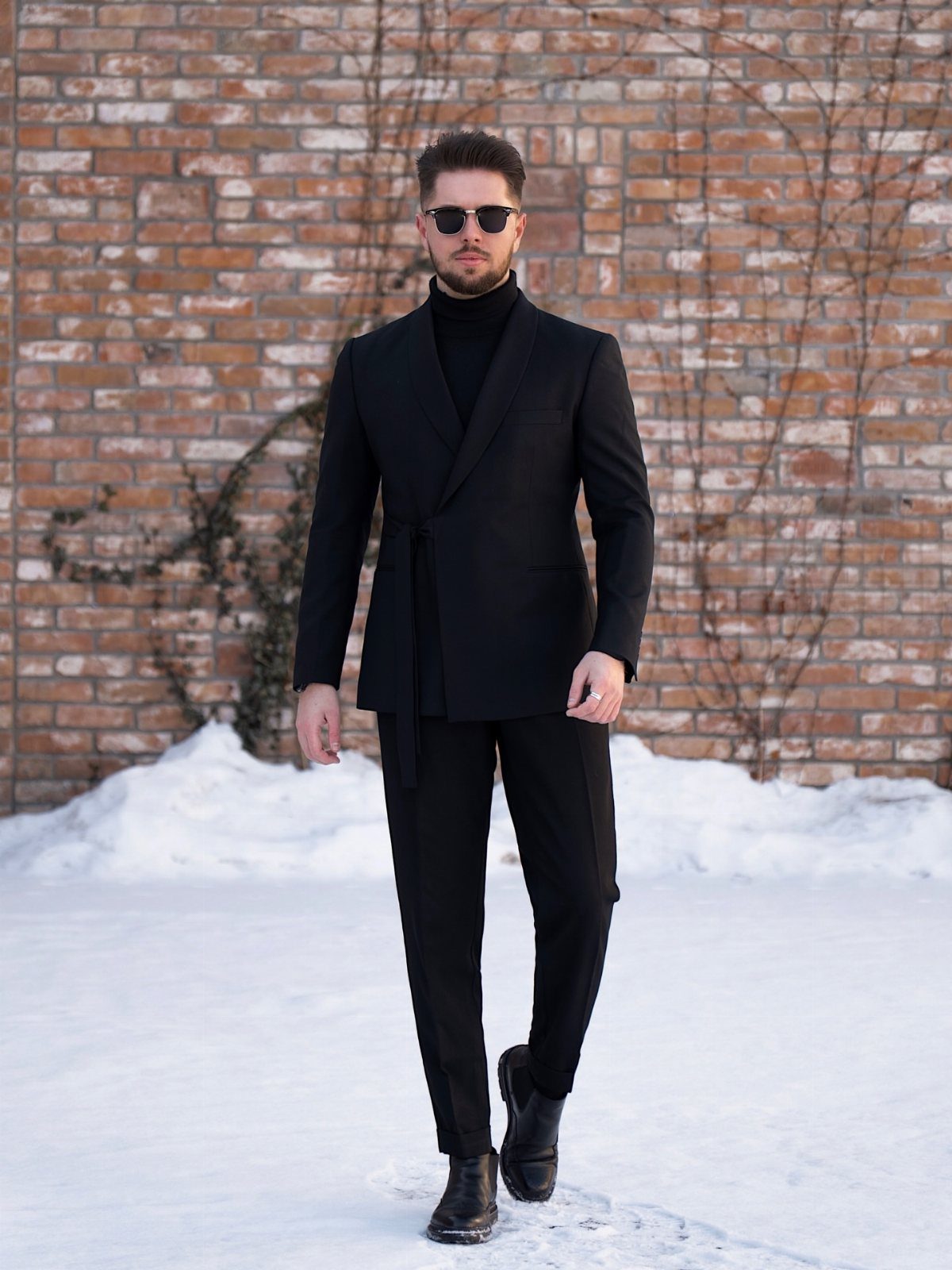 Man in an all black outfit including a blazer, turtleneck, and trousers standing in snow.