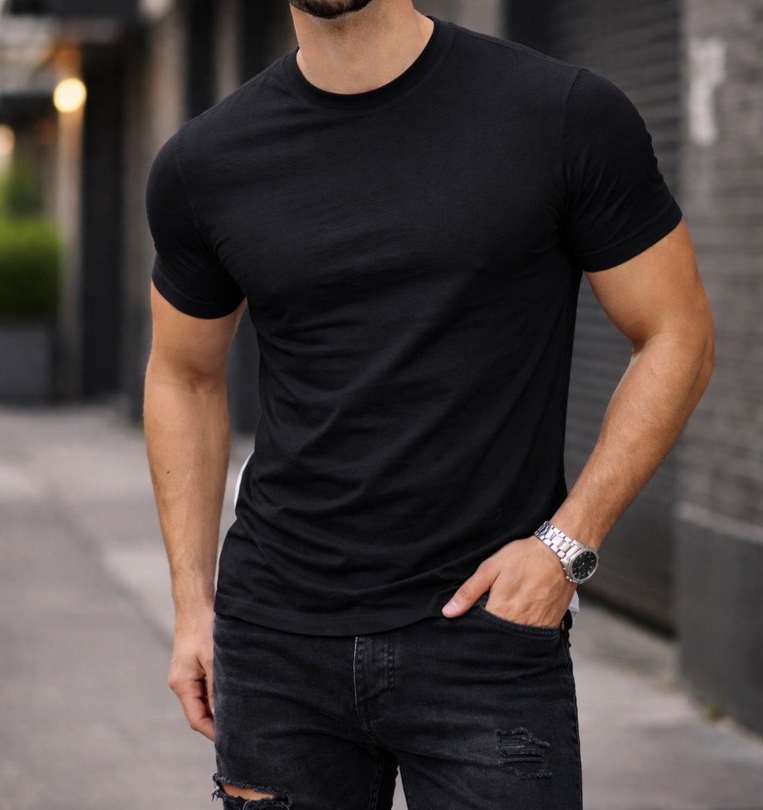 A muscular man wearing a black t-shirt and distressed black jeans standing on a street.