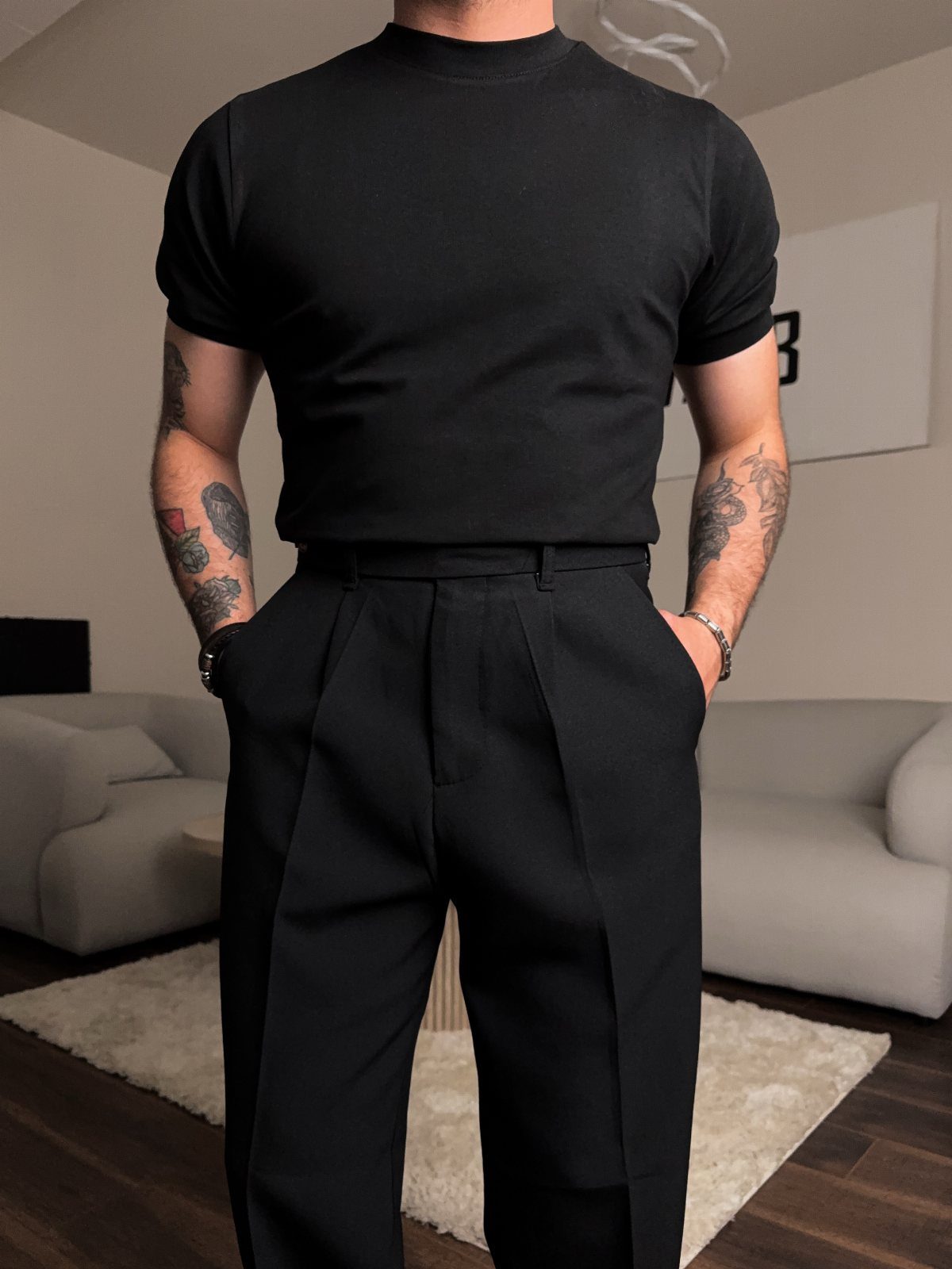 Man wearing a plain black t-shirt and black pleated tailored trousers standing in a room.
